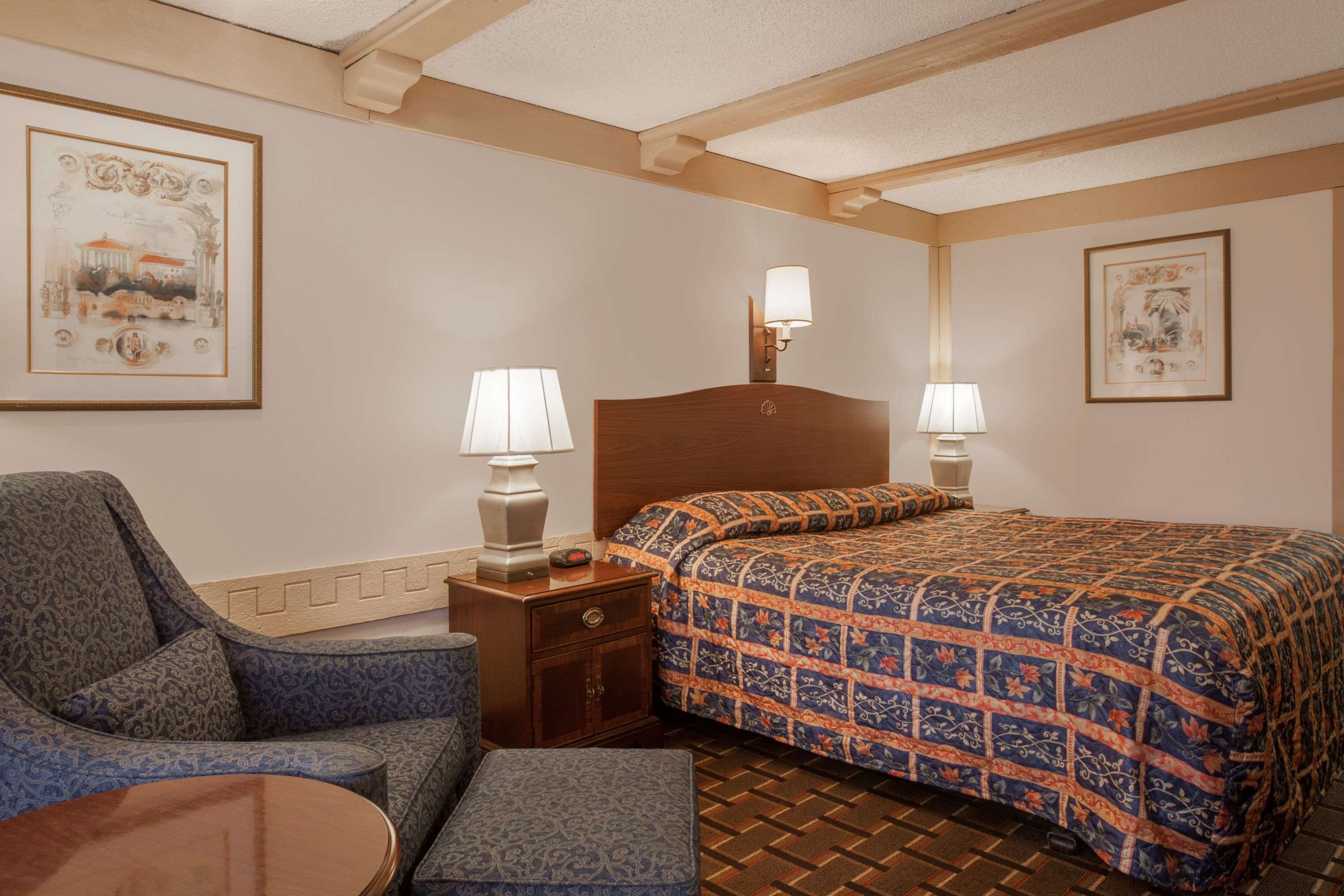 A hotel room with a bed, a chair, a small table, and two bedside lamps.