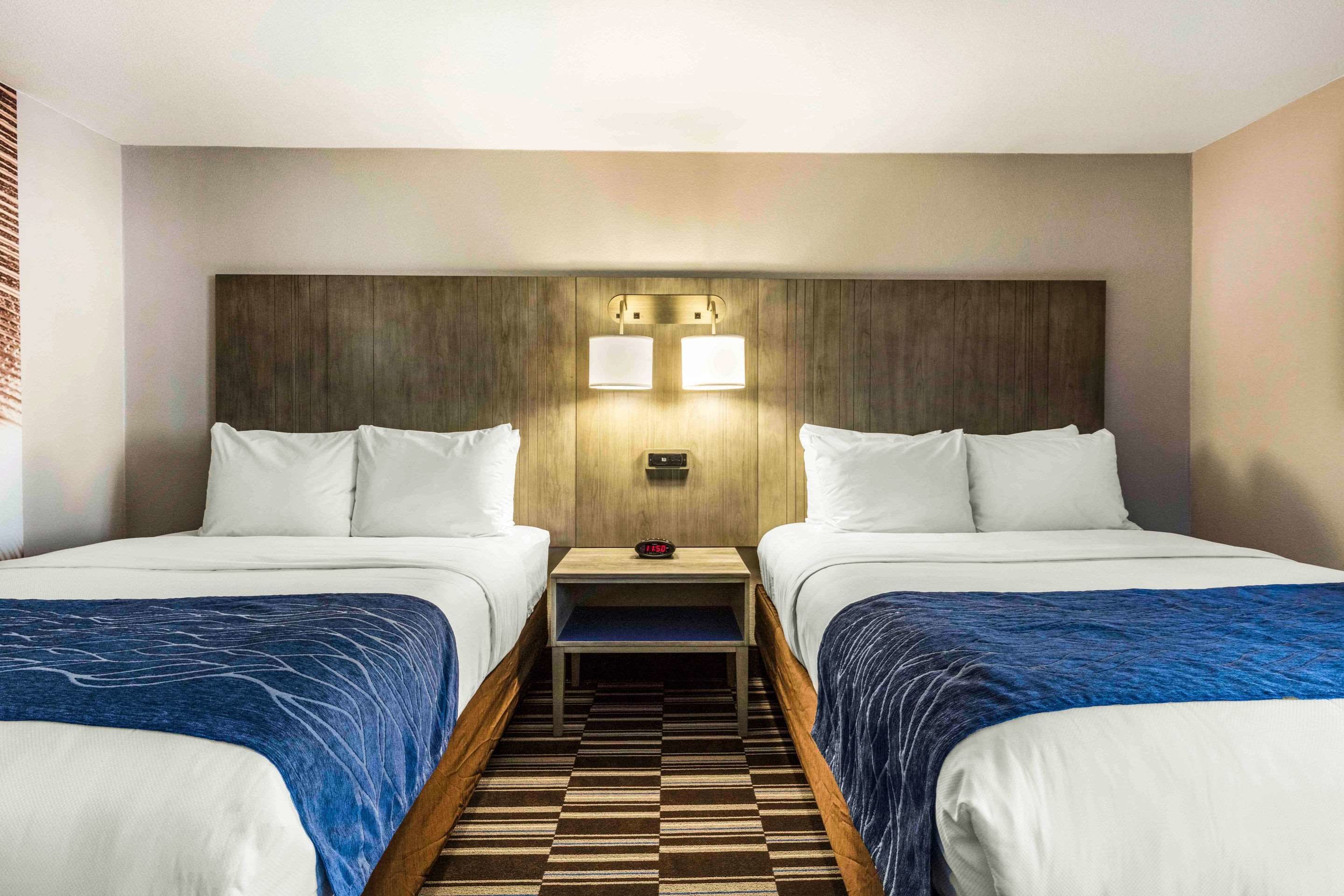 A hotel room with two beds, a wooden headboard, a nightstand with a clock, and a check-in station.