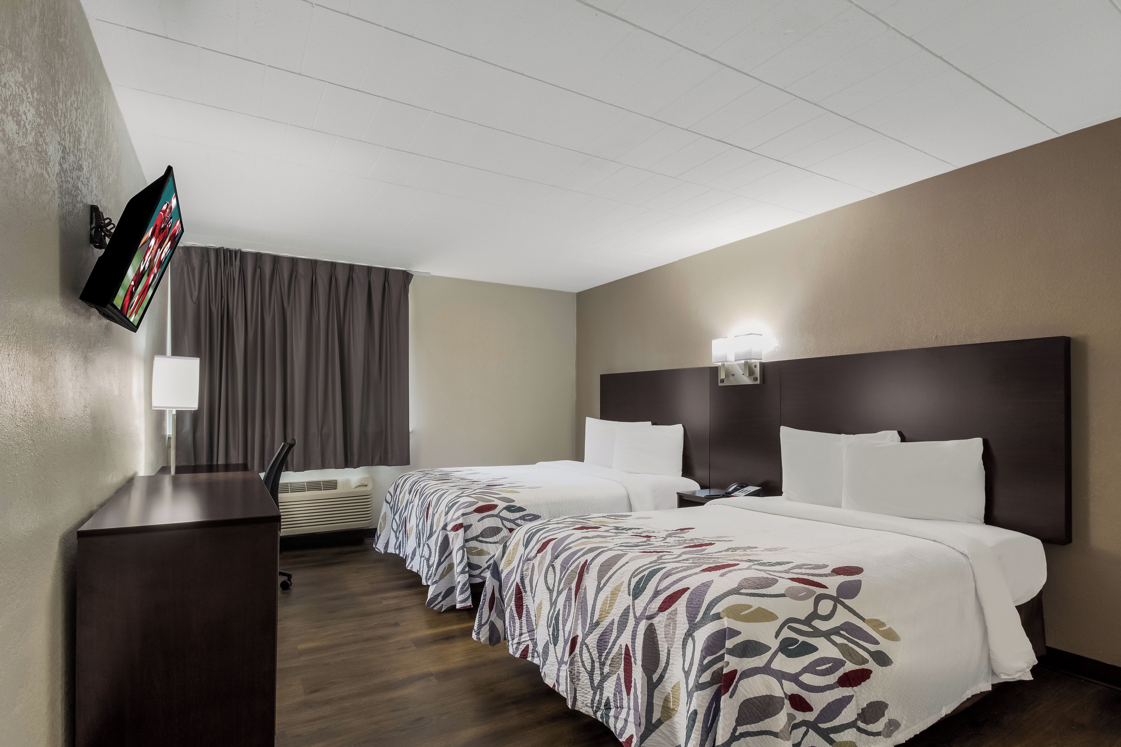 A hotel room with two beds, a flat-screen TV mounted on the wall, a desk, and a window with curtains.