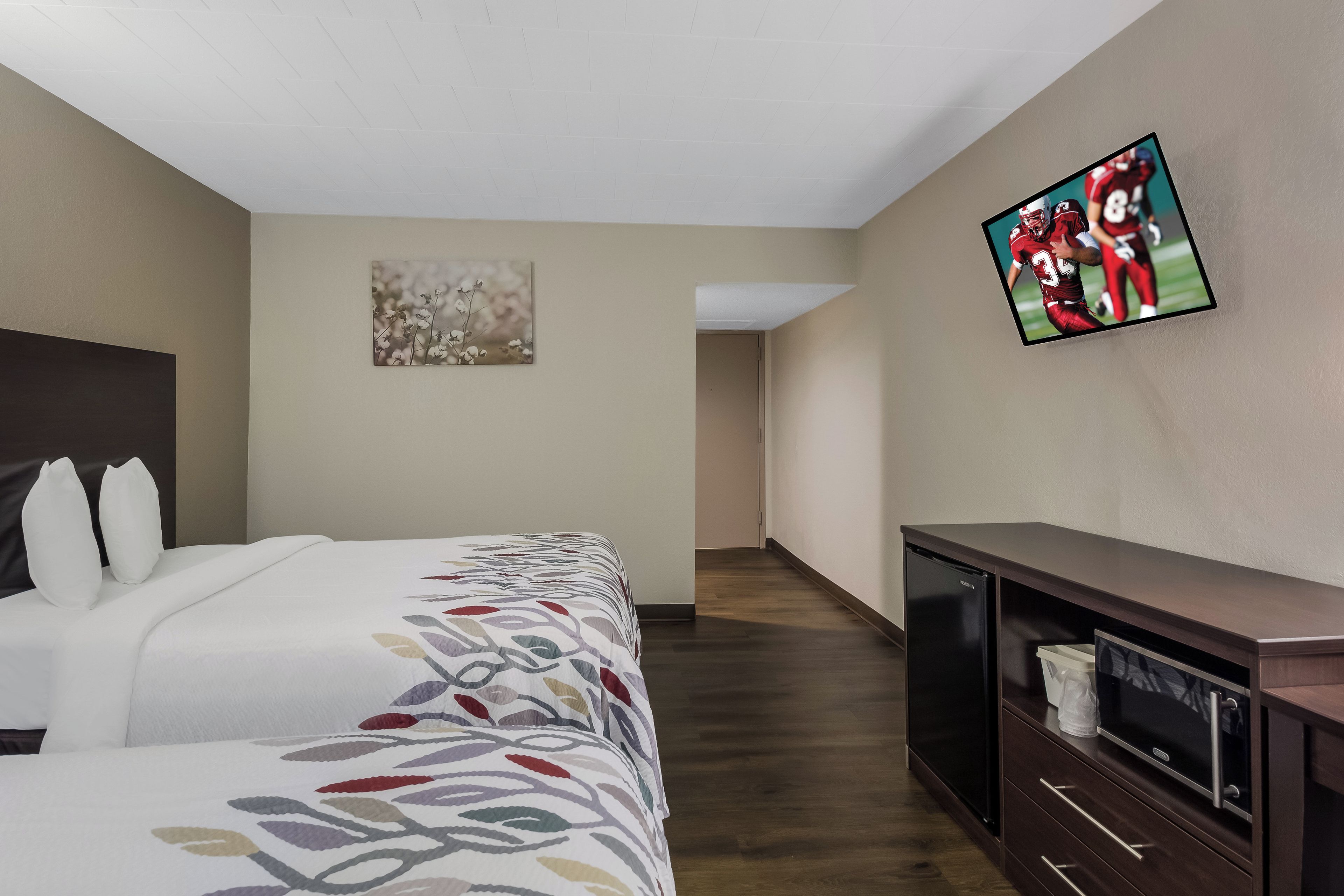 A hotel room with a bed, a flat-screen TV mounted on the wall, a small cabinet, and a door leading to another room.