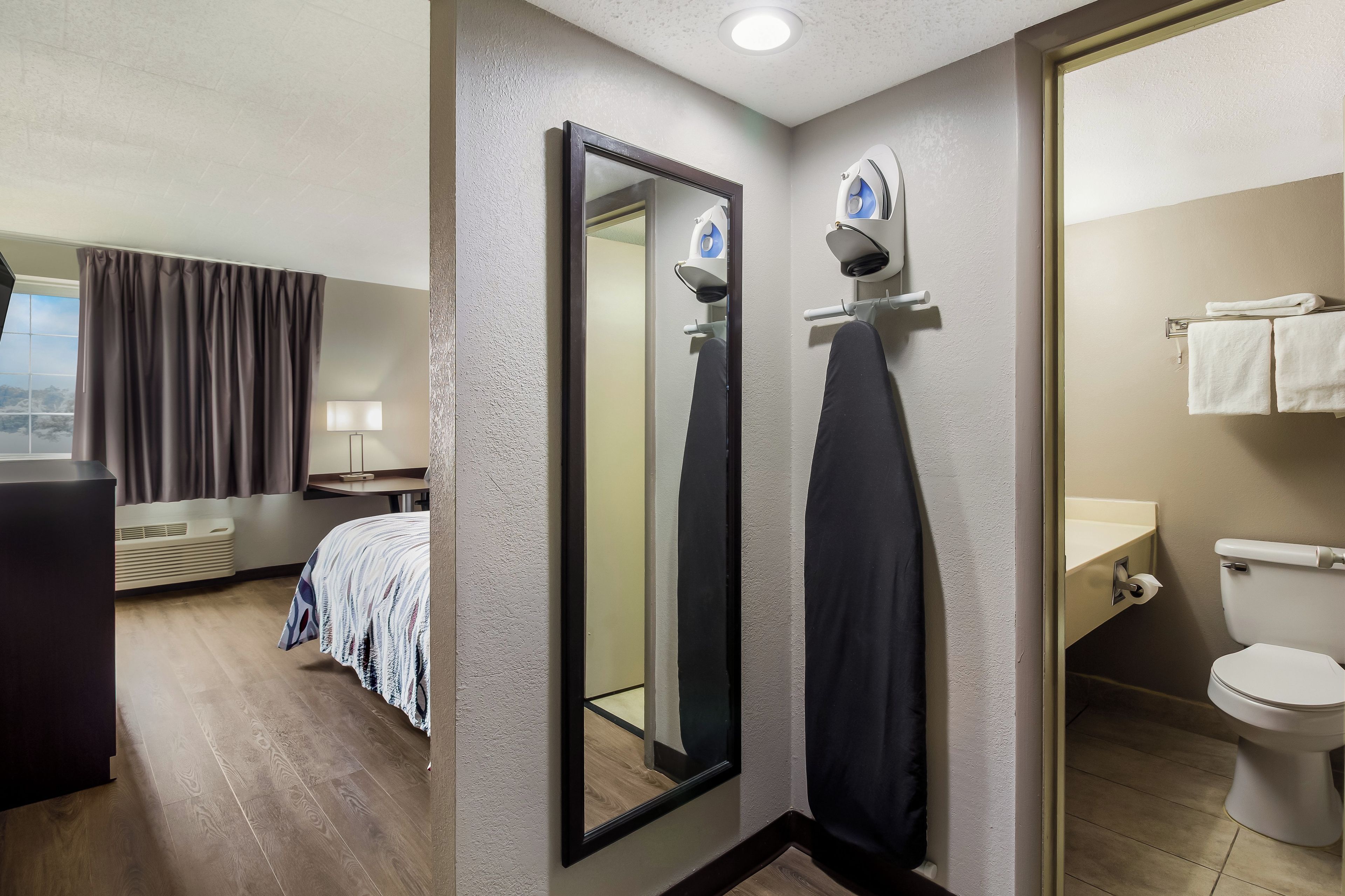 Una habitación de hotel con cama, espejo, baño con inodoro y ventana con cortinas.