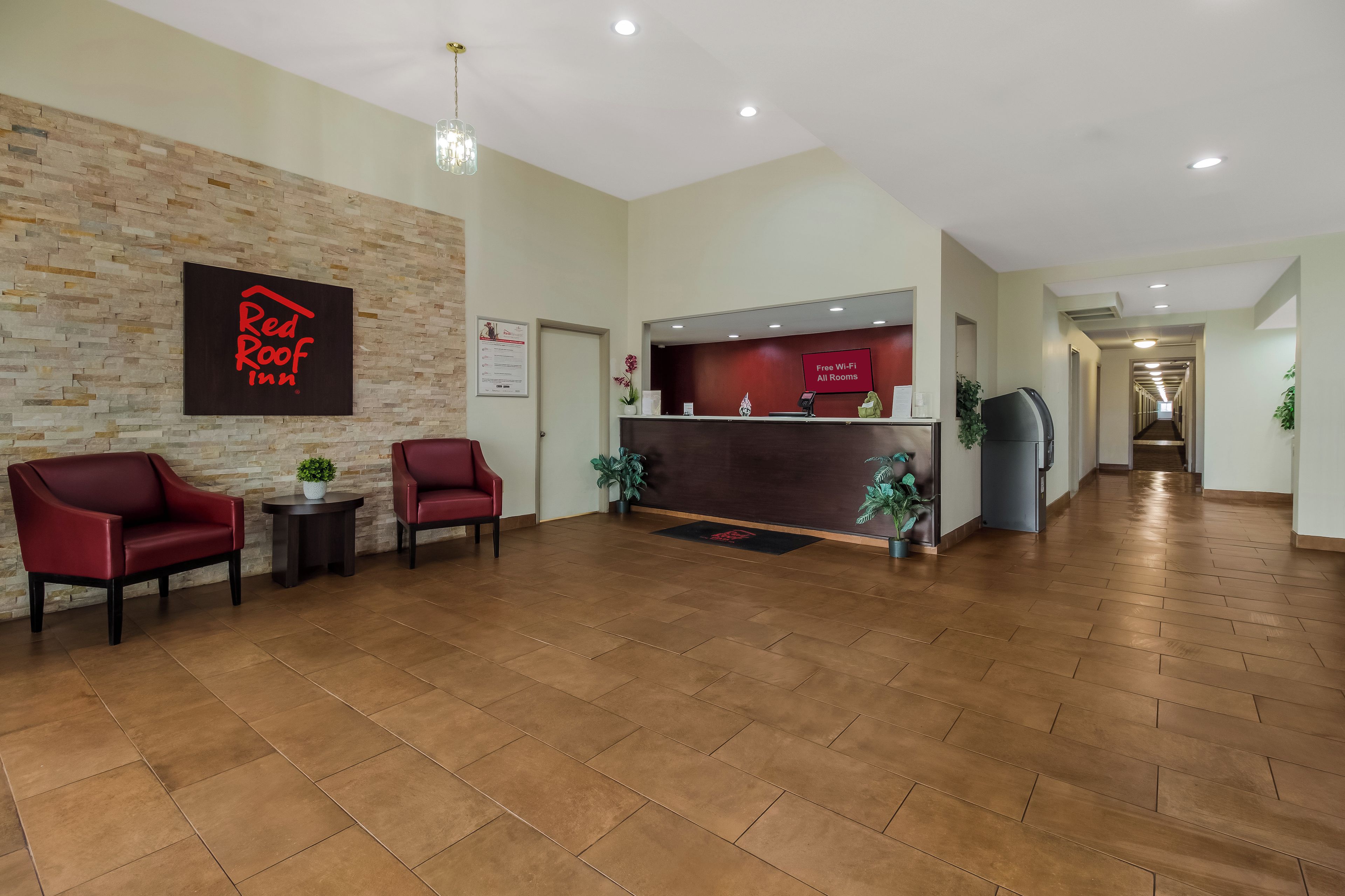 Zona de recepción de hotel con un letrero de Red Roof Inn, un mostrador de recepción, dos sillas rojas, una planta en maceta y un pasillo que conduce a otras habitaciones.