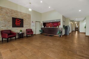 Ein Hotel-Empfangsbereich mit einem Red Roof Inn Schild, einem Empfangstresen, zwei roten Stühlen, einem Blumentopf und einem Gang zu weiteren Zimmern.