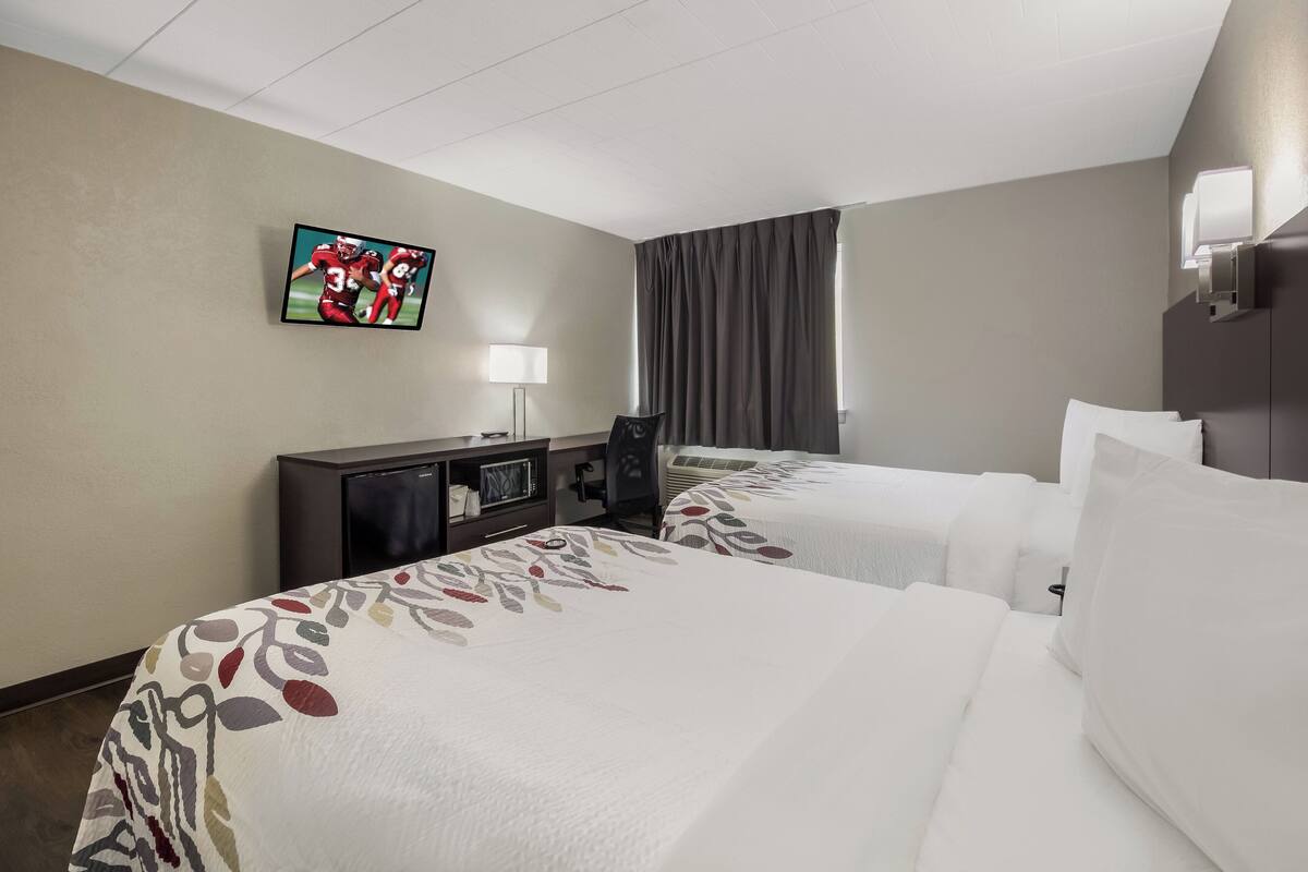Een hotelkamer met twee bedden, een televisie aan de muur, een bureau met een lamp en een raam met gordijnen.