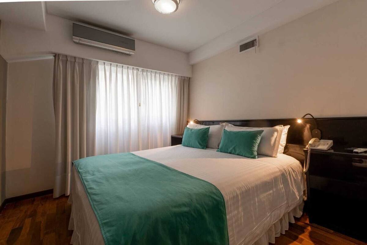 A hotel room with a bed, a bedside table with a lamp, a telephone, and a window with curtains.