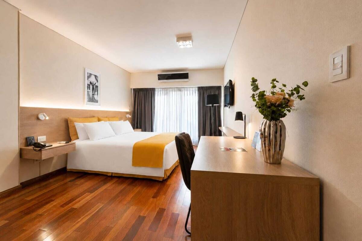 A hotel room with a bed, bedside table, desk, chair, and a vase with flowers.
