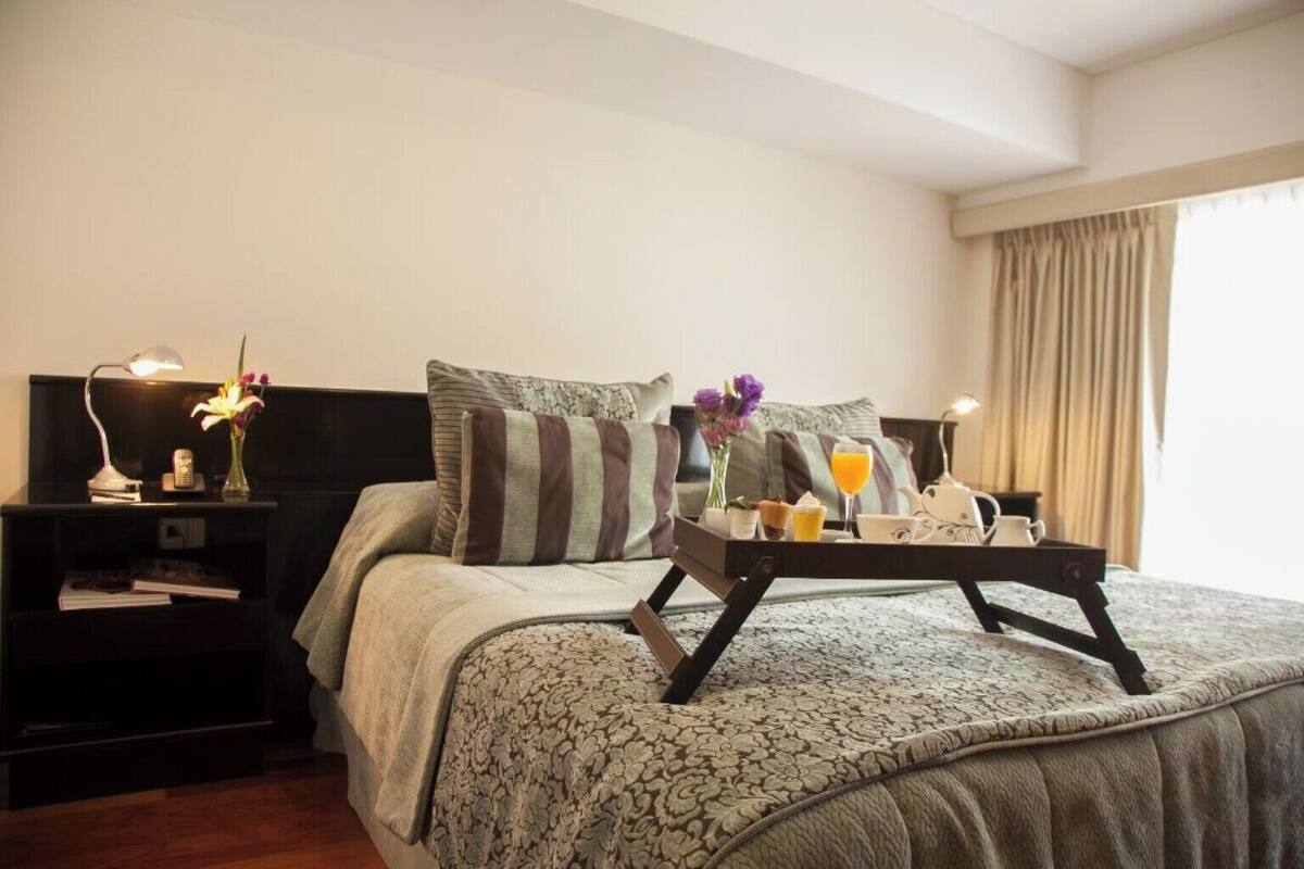 A hotel room with a bed, a bedside table with a lamp, a tray with breakfast items, and a window with curtains.