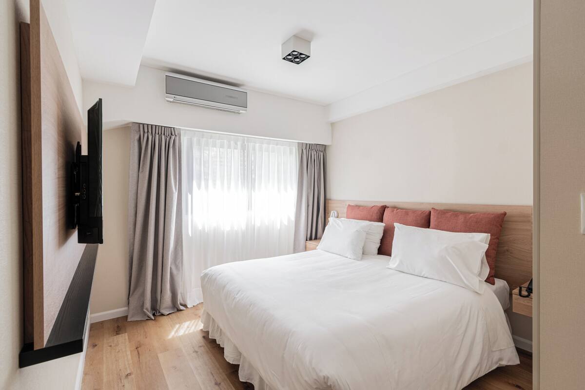 A hotel room with a bed, curtains, a mounted TV, and an air conditioning unit.