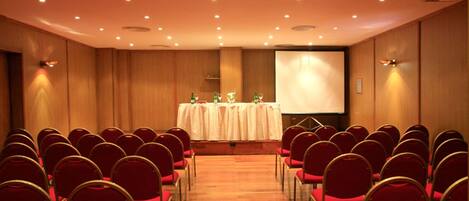 A conference room with rows of red chairs, a stage with a projector screen, and a table with water bottles.
