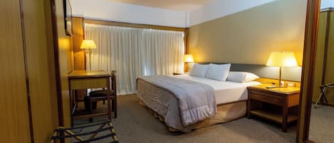 A hotel room with a bed, bedside tables, a desk, a chair, a mirror, and a window with curtains.