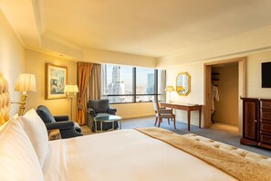 A hotel room with a large bed, a desk, a chair, and a view of the cityscape through the window.