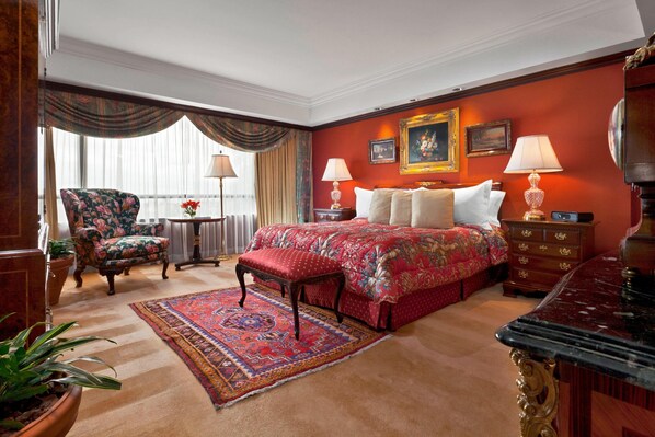 Une chambre d’hôtel avec un grand lit, un fauteuil à motifs floraux, une commode en bois et une moquette rouge.