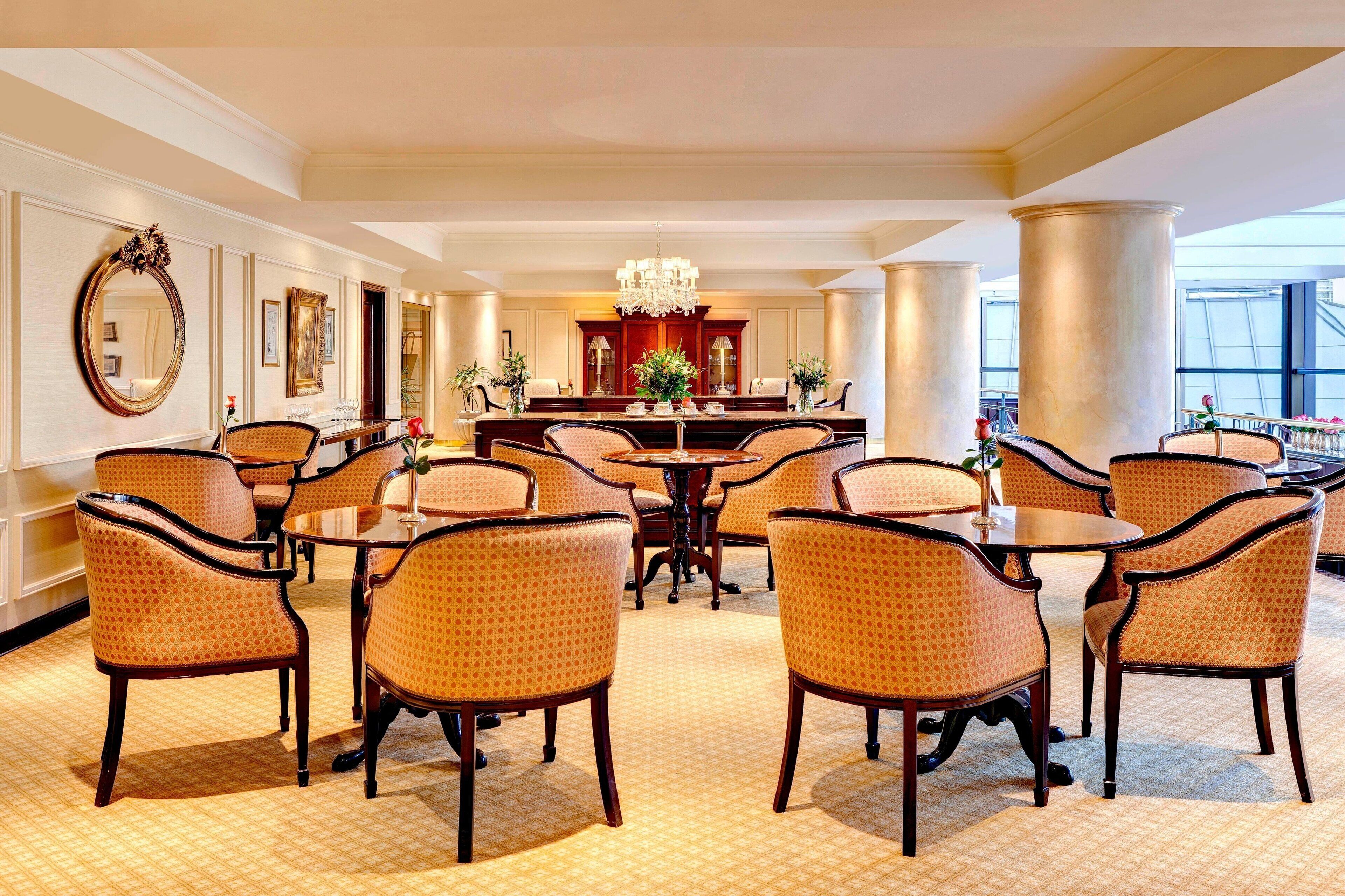 Un comedor amplio con mesas y sillas redondas, un candelabro central y ventanas grandes.