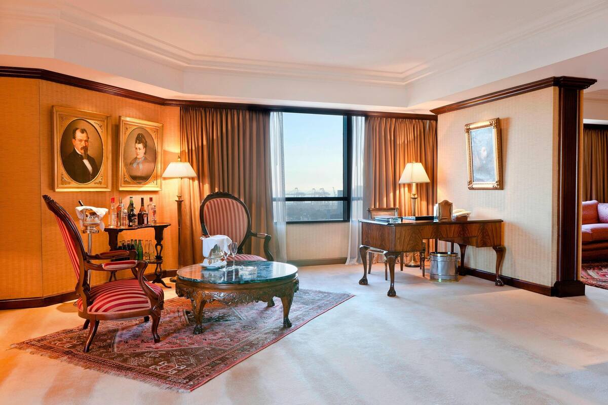 A hotel room with a large window, a piano, a dining table, a striped armchair, and a coffee table.