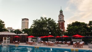 Zwembad met rode parasols en ligbedden, een klokkentoren, en moderne gebouwen op de achtergrond.