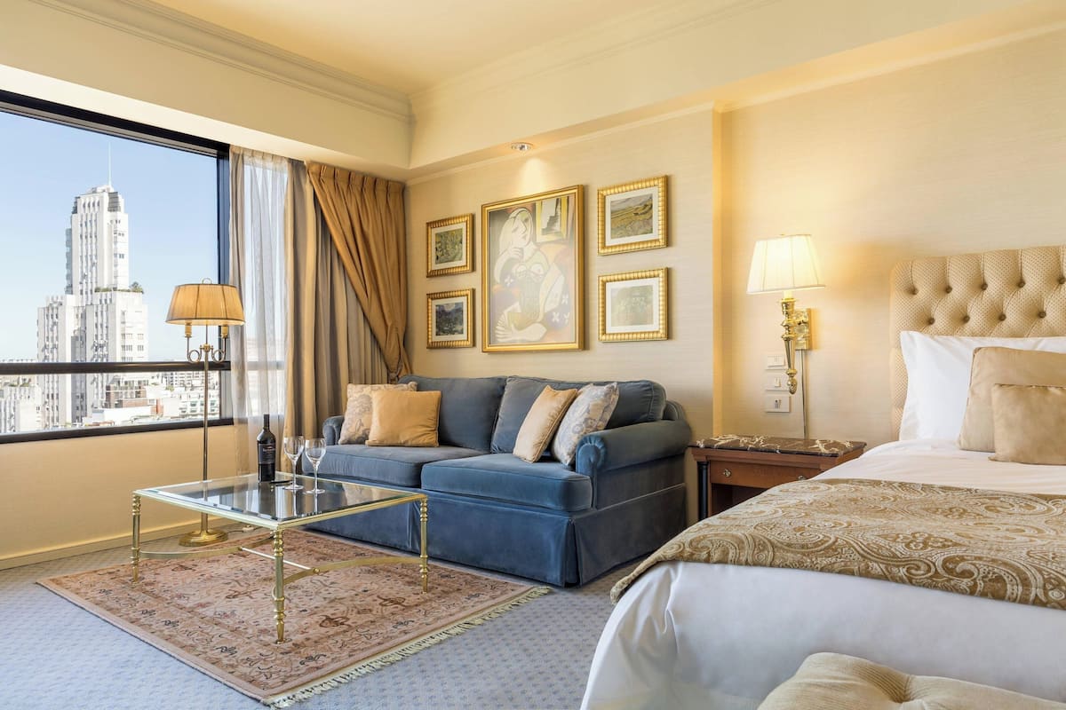 A hotel room with a bed, a sofa, a glass coffee table, and a view of a cityscape.