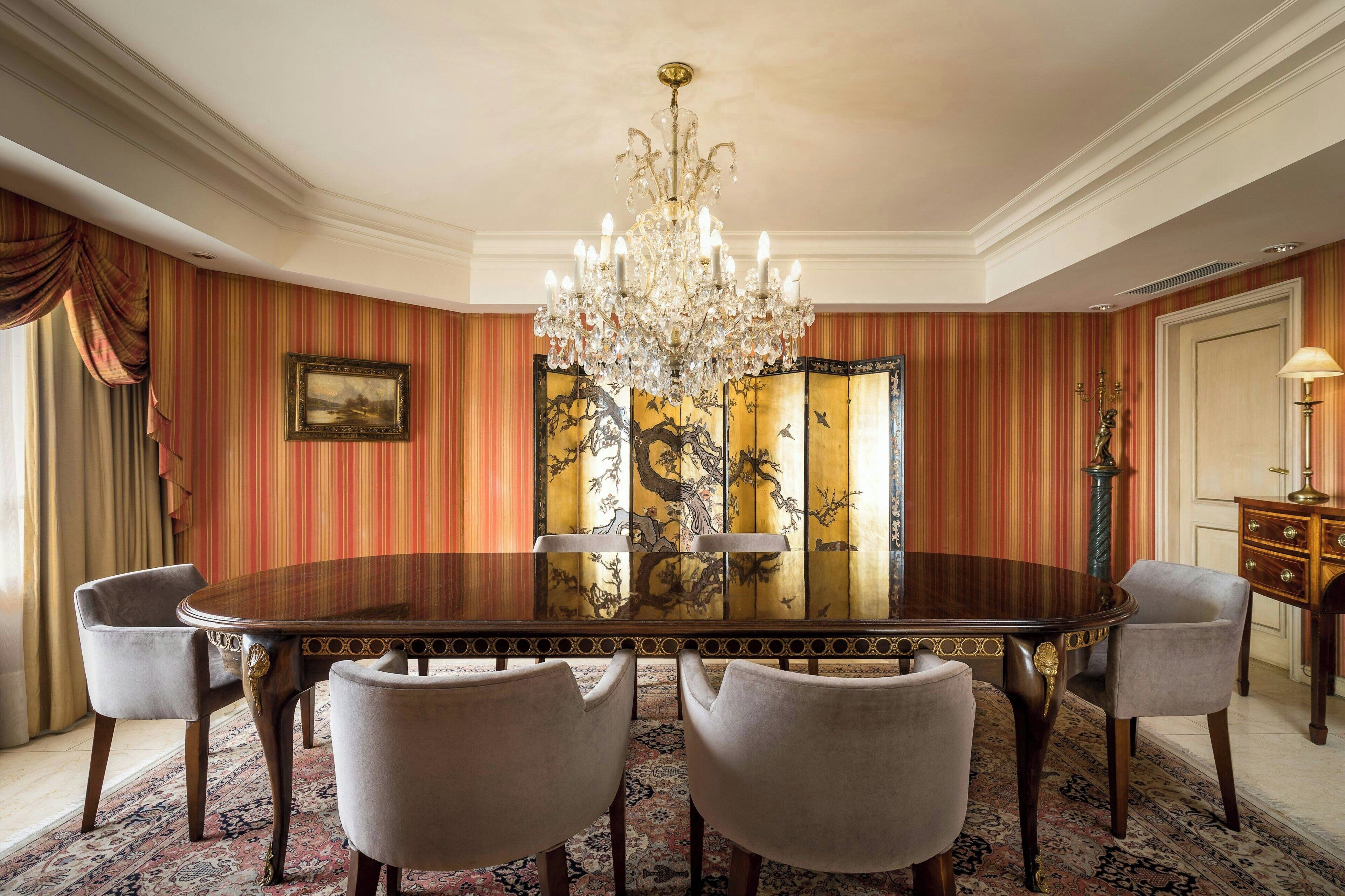 Un comedor con una mesa redonda, seis sillas, una araña, un biombo de estilo asiático tradicional y un mueble de madera con cajones.