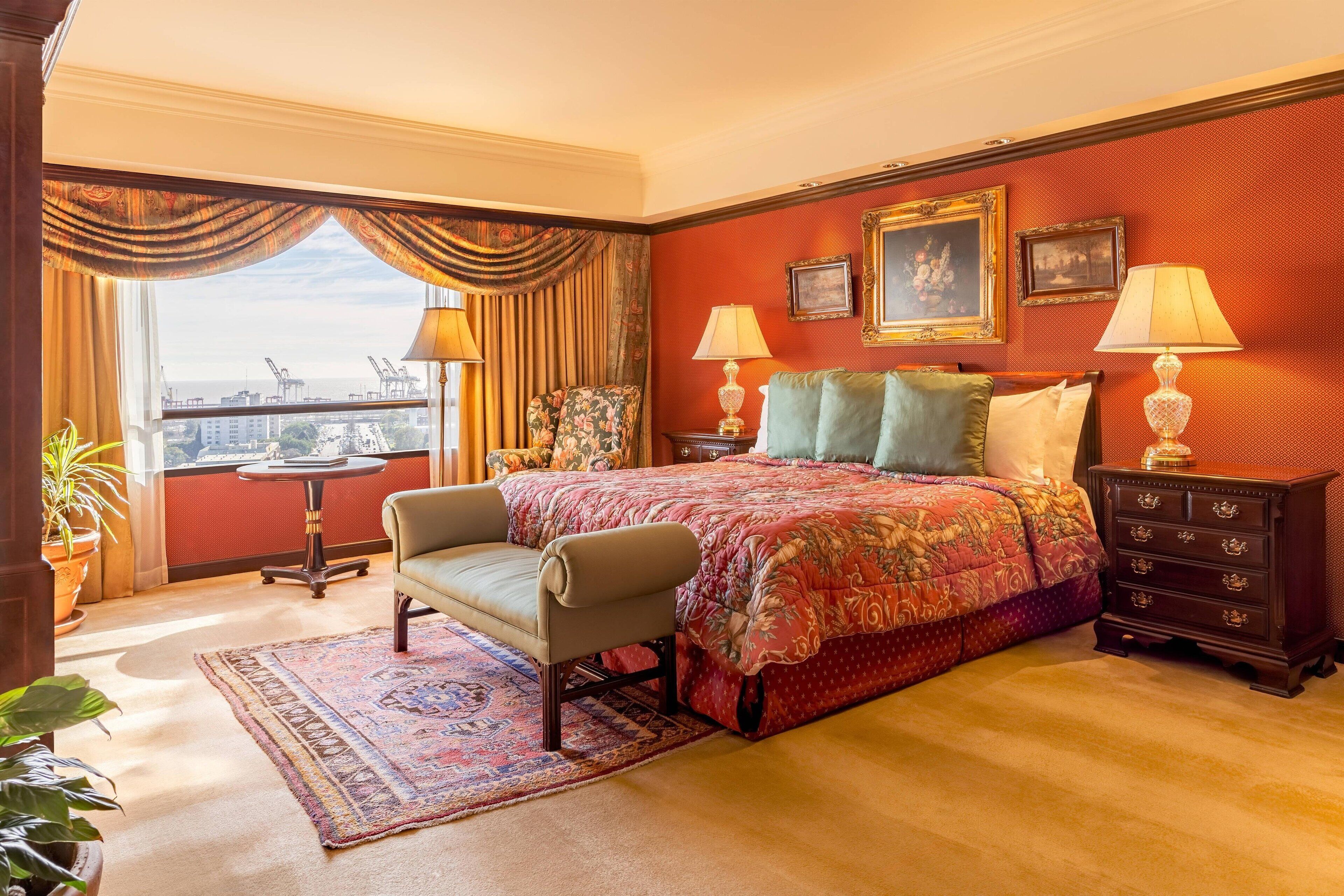 A hotel room with a large bed, two bedside tables with lamps, a chair, a small table, and a view of the city through a window with curtains.