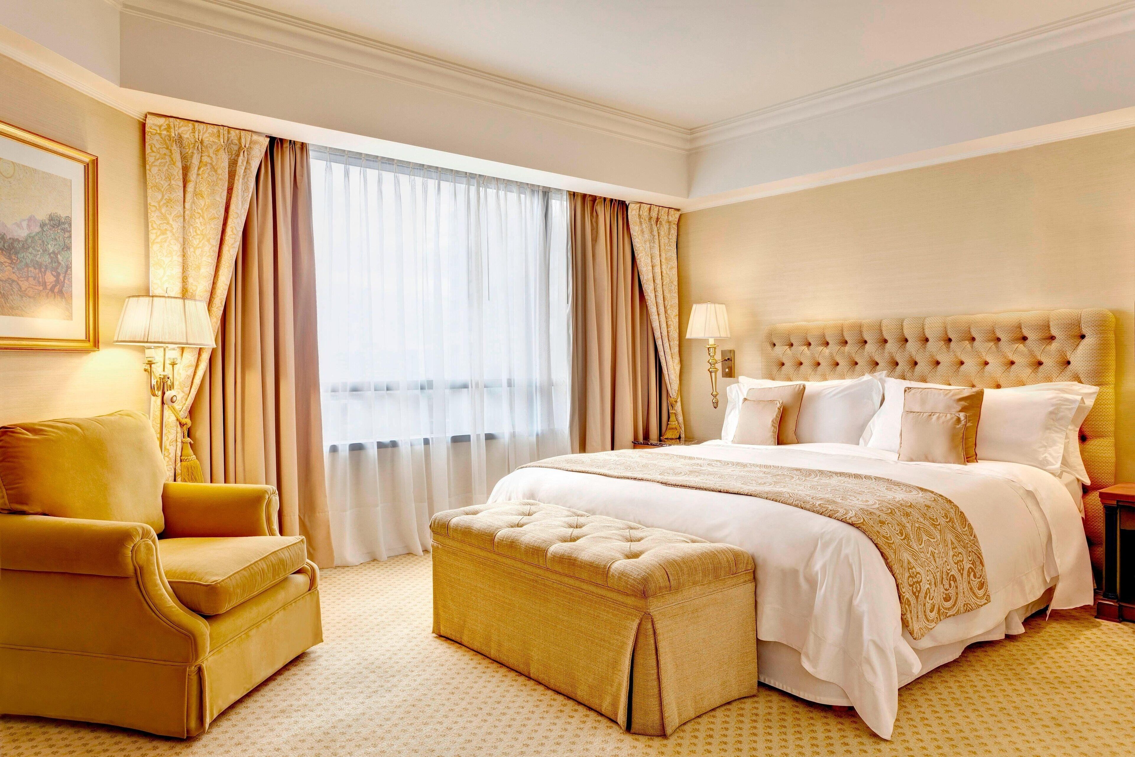 A hotel room with a large bed, a gold armchair, a small ottoman, and a painting on the wall.