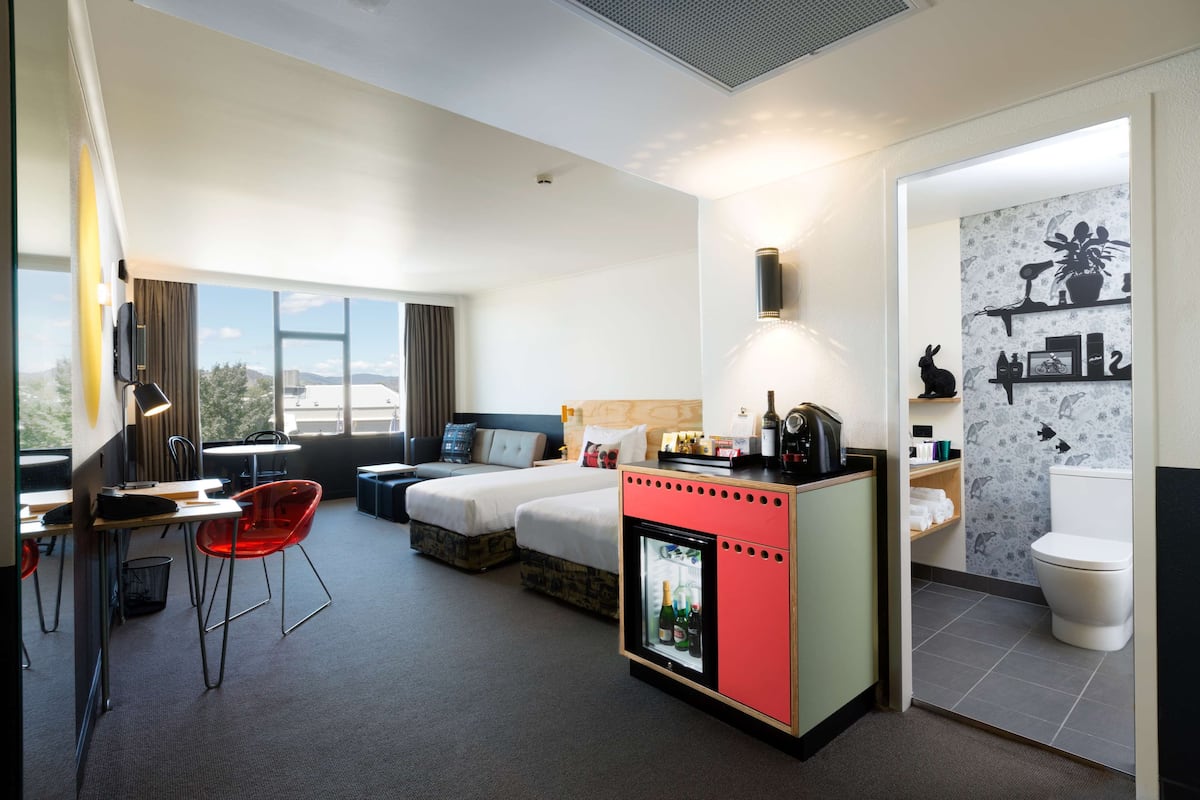 A hotel room with a bed, a desk with a chair, a mini-fridge, and a bathroom with a toilet and shelves.
