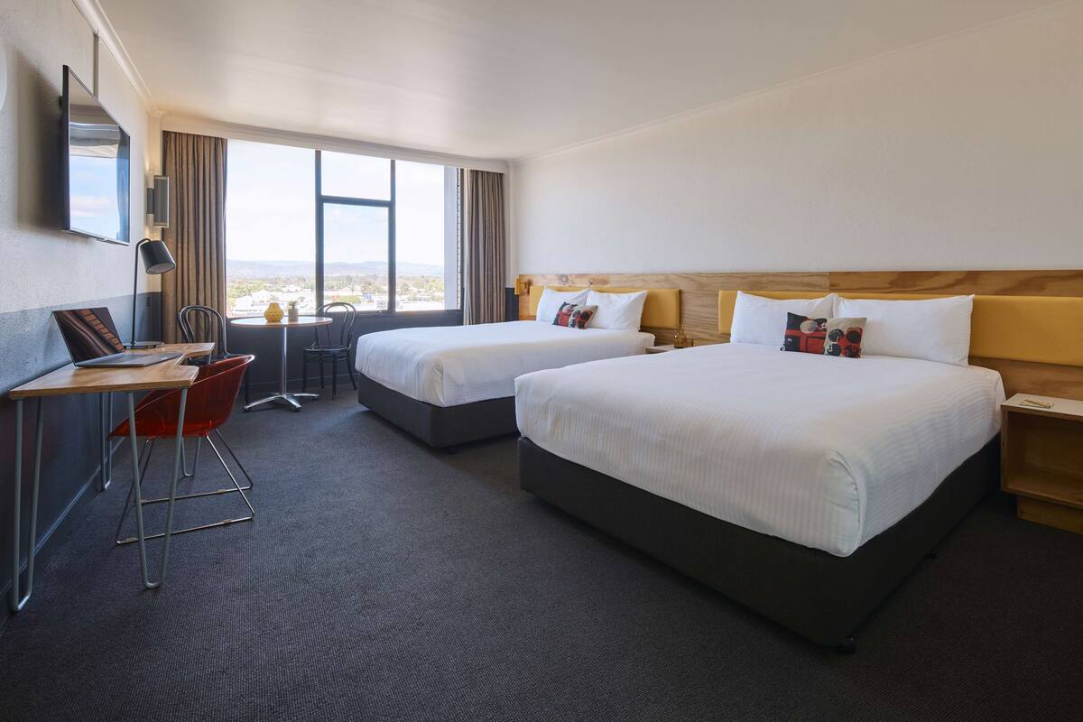 A hotel room with two beds, a desk with a chair, a TV mounted on the wall, and a view of the city.
