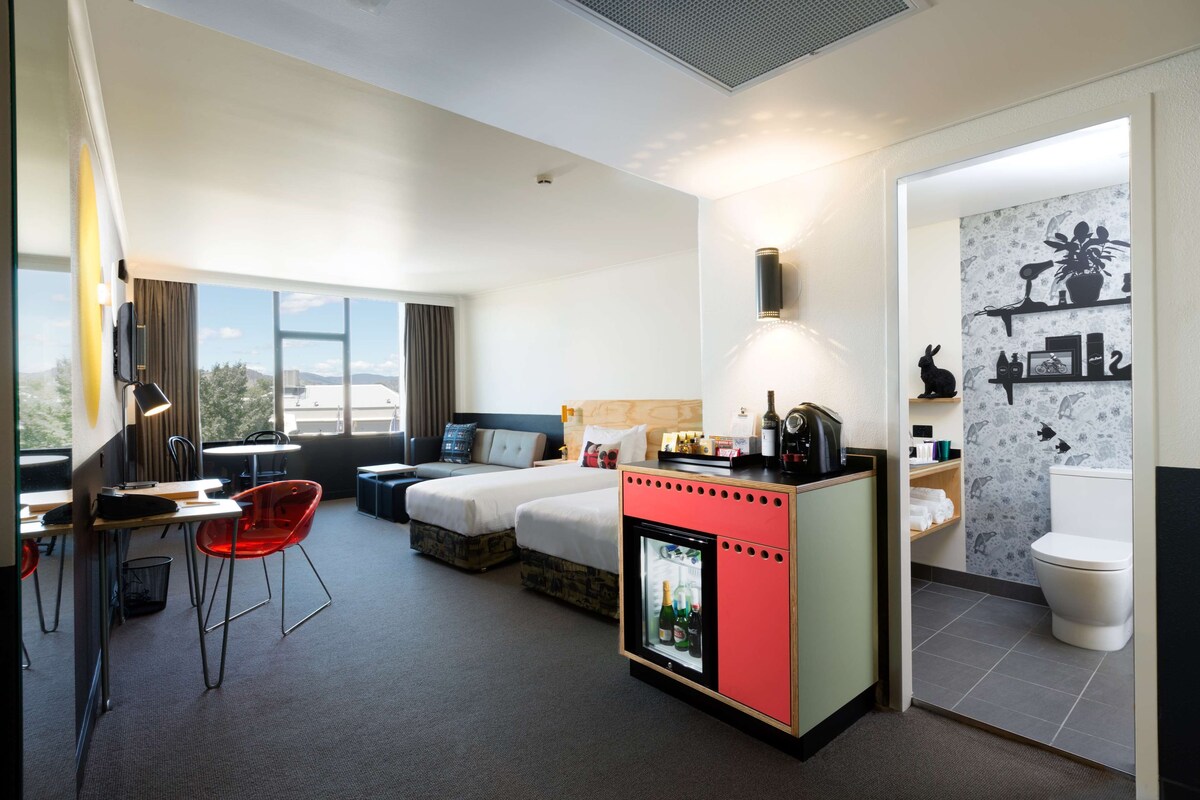 Habitación de hotel con cama, escritorio con silla, minibar, baño con inodoro y estanterías, y vista al exterior.