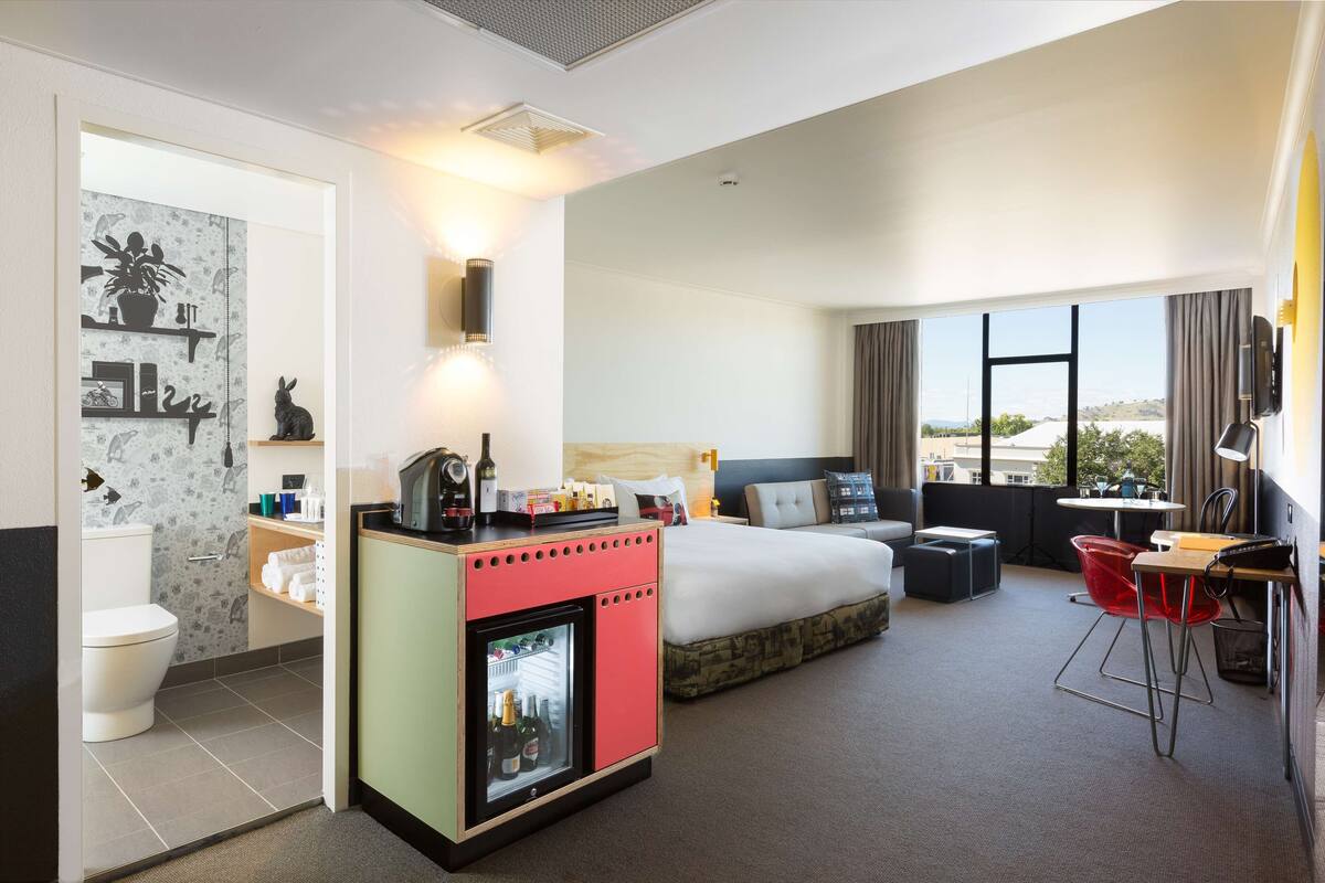 A hotel room with a bed, a small kitchenette, a seating area with a table and chairs, a TV, and a large window with a view.