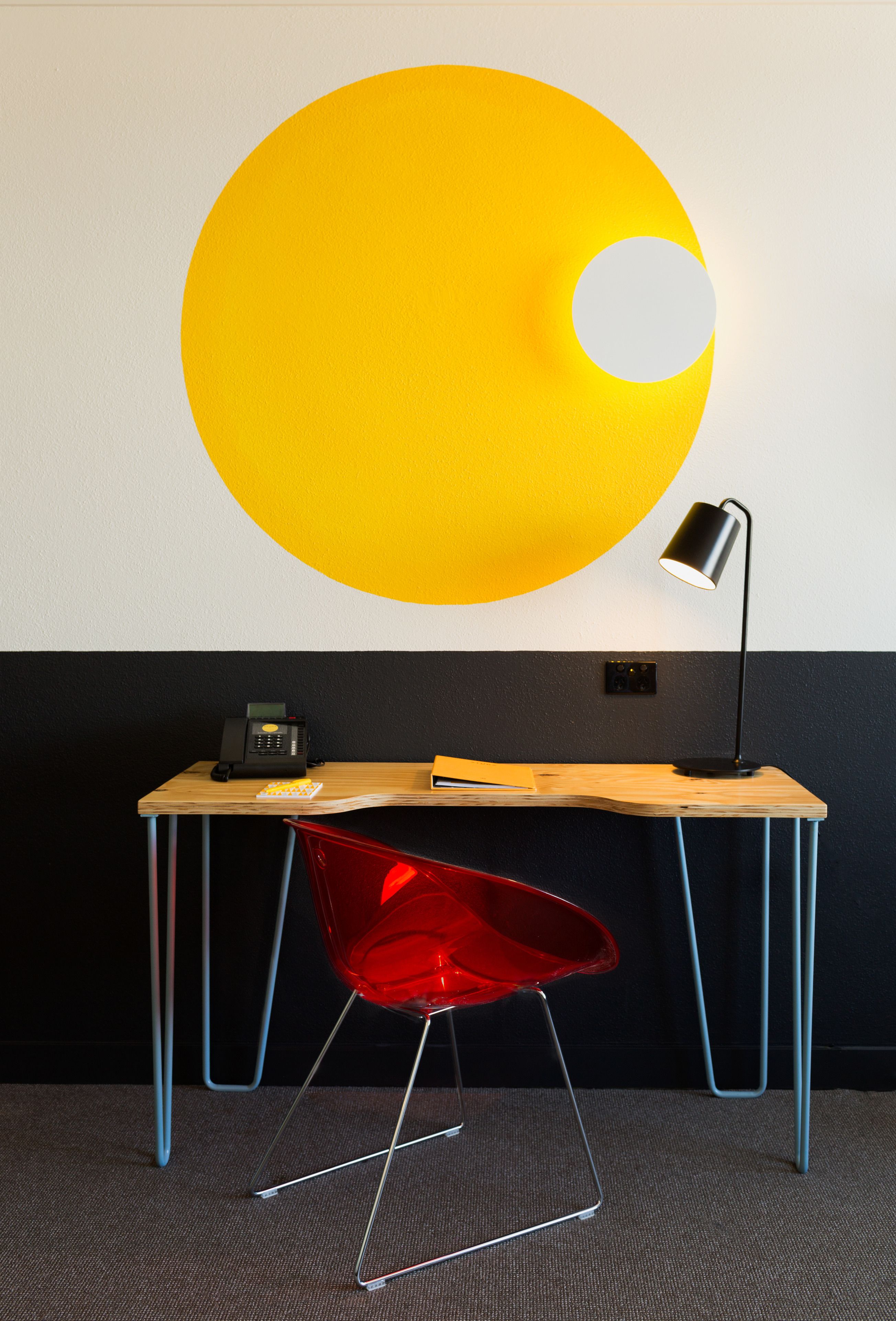 Uma mesa com uma luminária, uma cadeira vermelha e um grande adorno amarelo na parede.