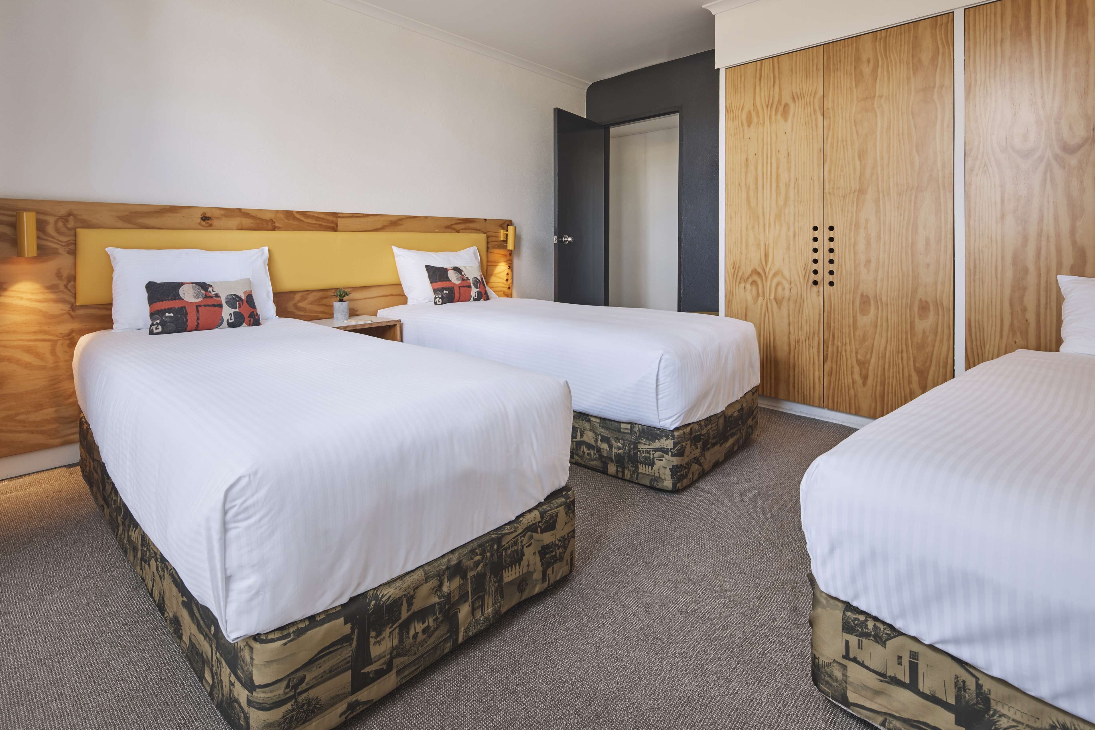 Une chambre d’hôtel avec deux lits simples, une tête de lit en bois, une armoire et une petite table de chevet avec une lampe.