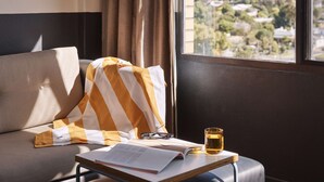 A room with a window, a sofa, a striped blanket, a pair of glasses, a glass of amber liquid, and a book on a small table. - Atura Albury (Albury)