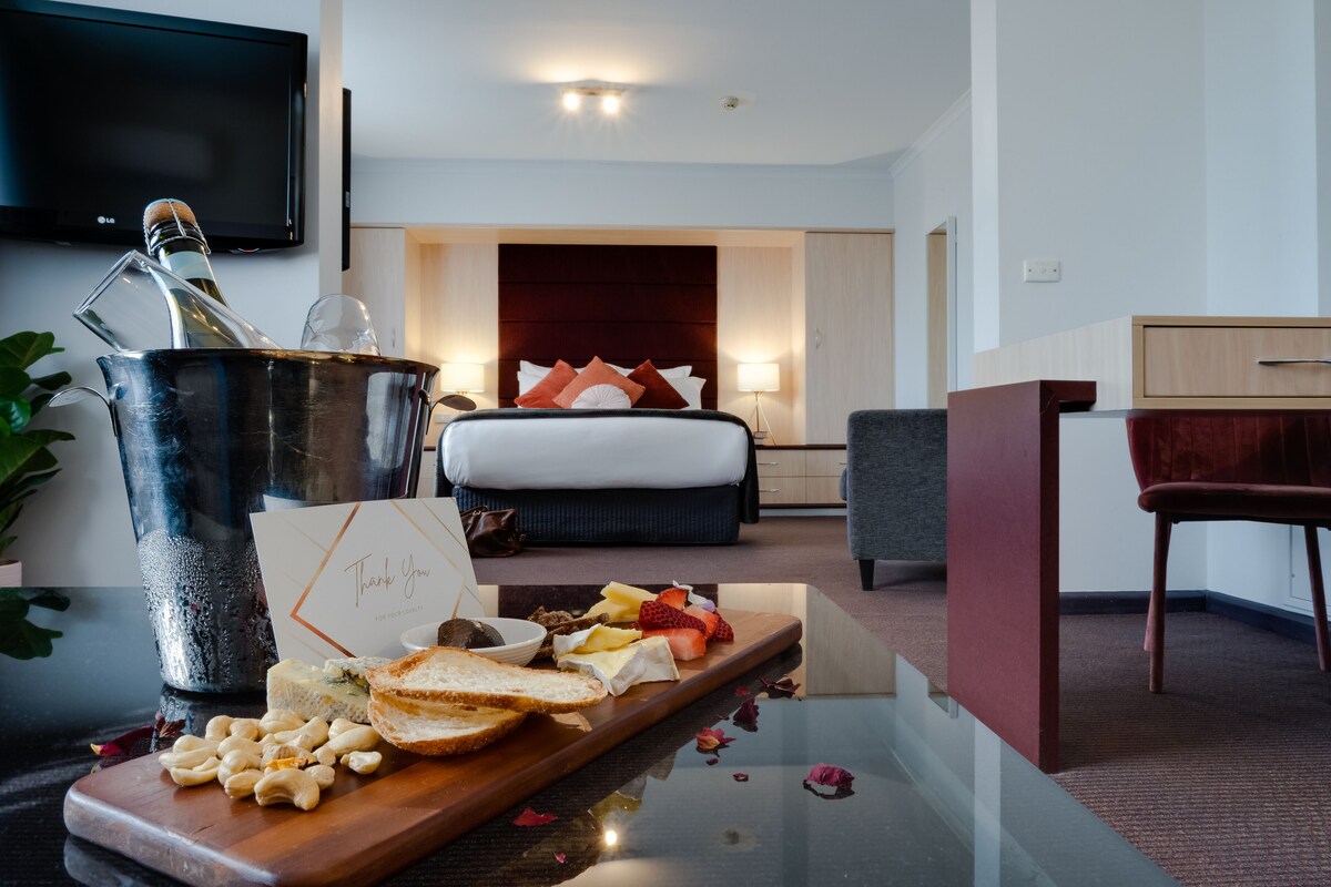 A hotel room with a bed, bedside table, a desk, a TV, and a breakfast tray with food and a bottle of champagne.