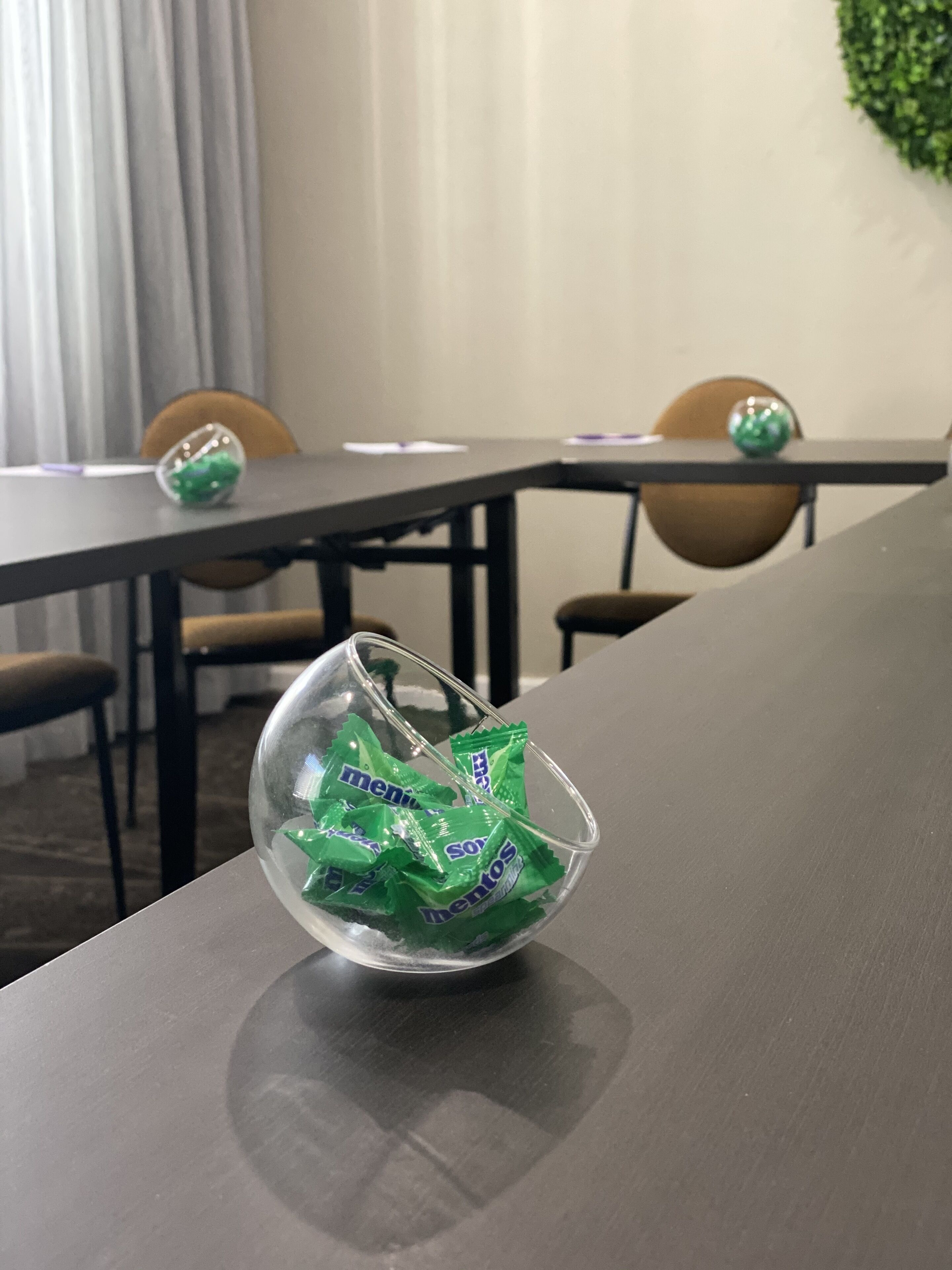 Ein Konferenztisch mit einer klaren Glasvase, die mit grünen Beuteln gefüllt ist, ein grauer Vorhang und eine Zimmerpflanze im Hintergrund.