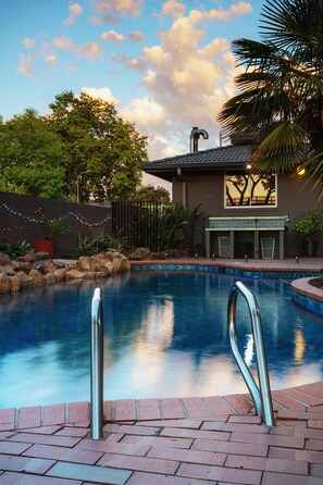 A swimming pool with metal ladders, surrounded by a brick patio, a house with a fireplace, and a view of trees and a sunset. - Mercure Albury (Albury)