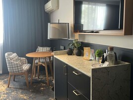 Habitación de hotel compacta con sofá, una pequeña mesa redonda, televisión montada en la pared, minibar y una planta en maceta.
