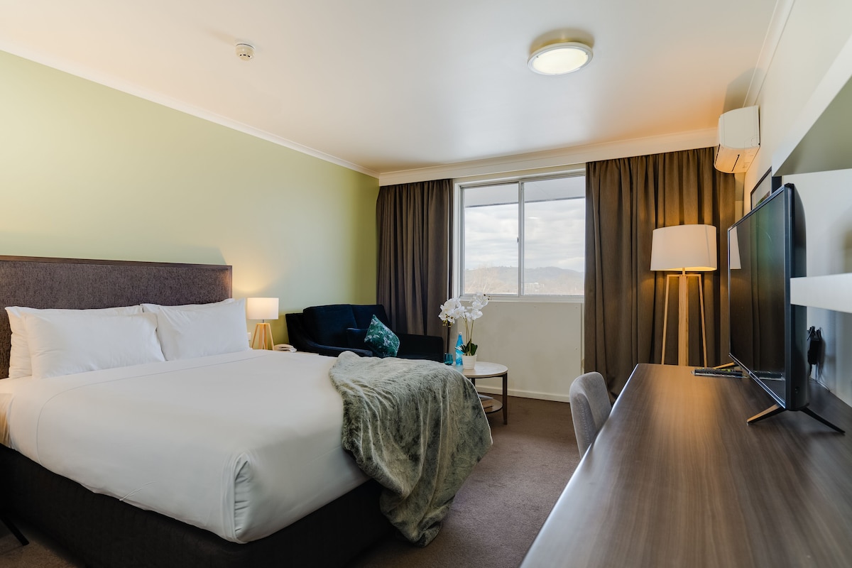 A hotel room with a large bed, a desk, a chair, a TV, and a window with curtains.