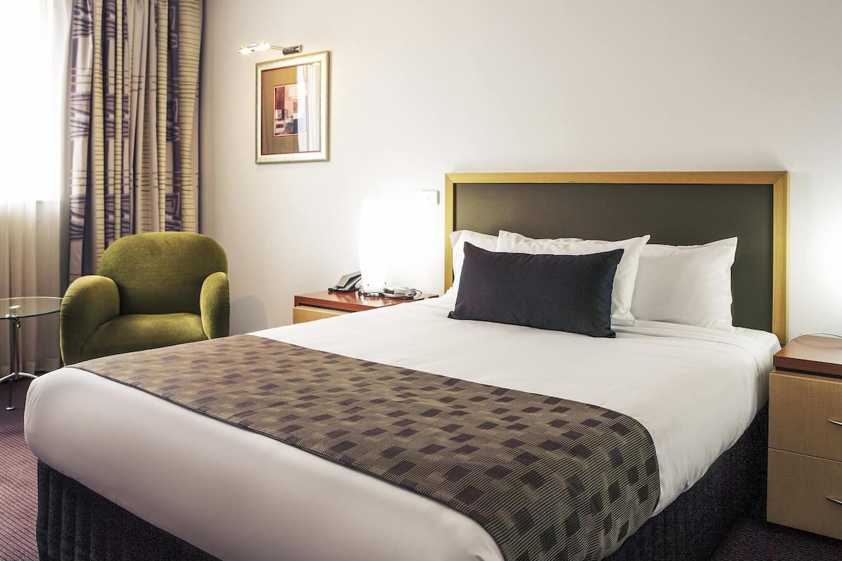 A hotel room with a bed, a green armchair, a bedside table with a lamp, and a framed picture on the wall.