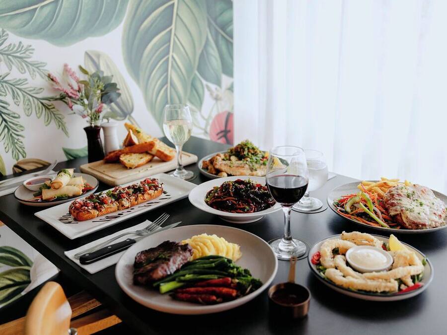 A dining table with various dishes and drinks, set against a backdrop of a large botanical print.