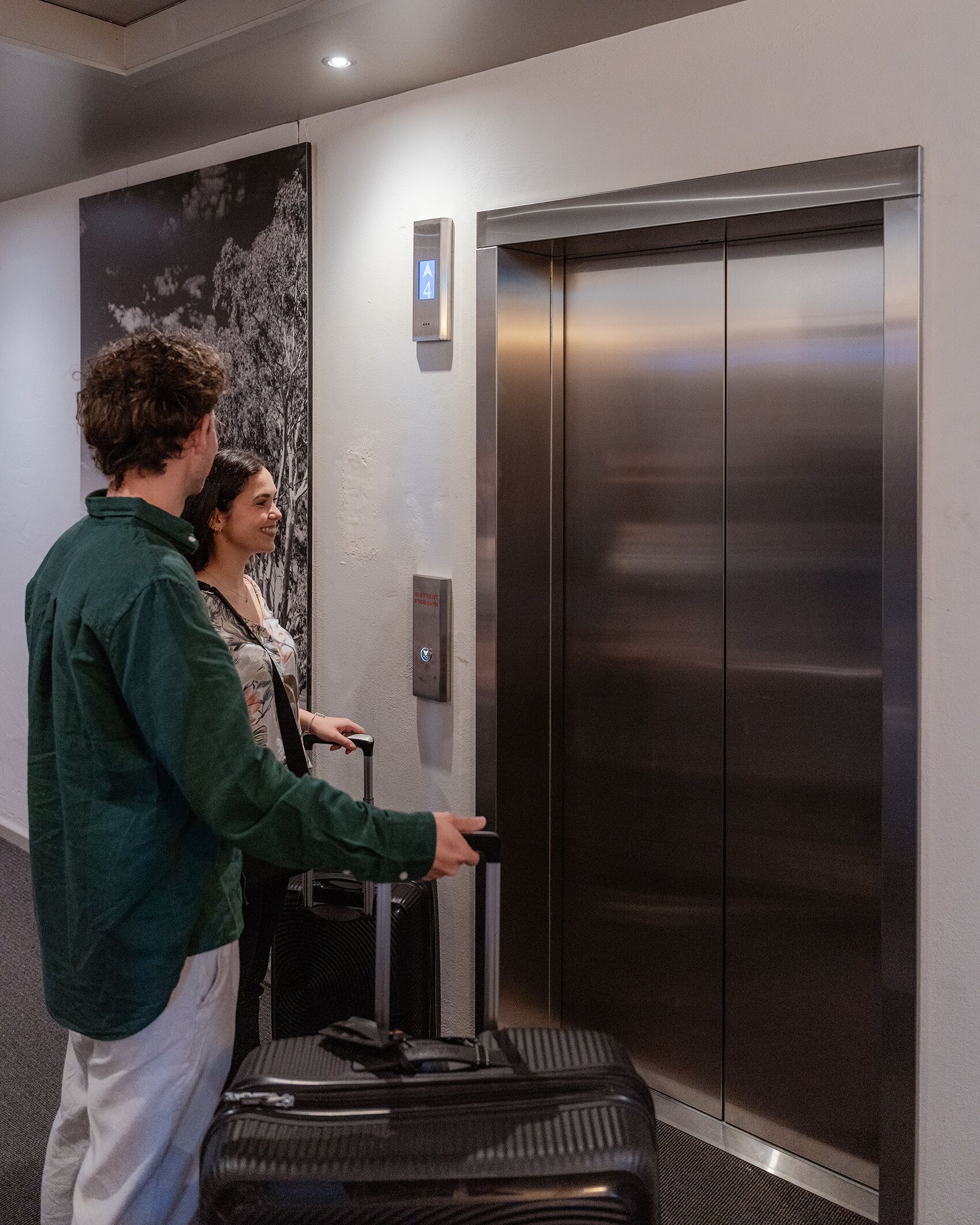 A man and a woman standing next to a wall-mounted elevator in a hotel lobby.