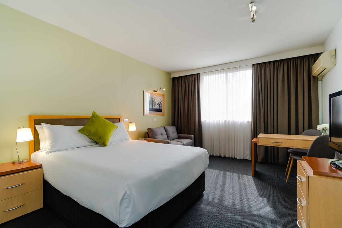A hotel room with a large bed, a desk, a chair, a TV, and a window with curtains.