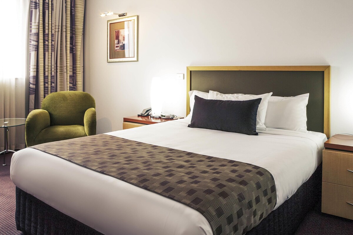 Una habitación de hotel con una cama, un sillón verde, una mesita de noche con una lámpara y un cuadro en la pared.