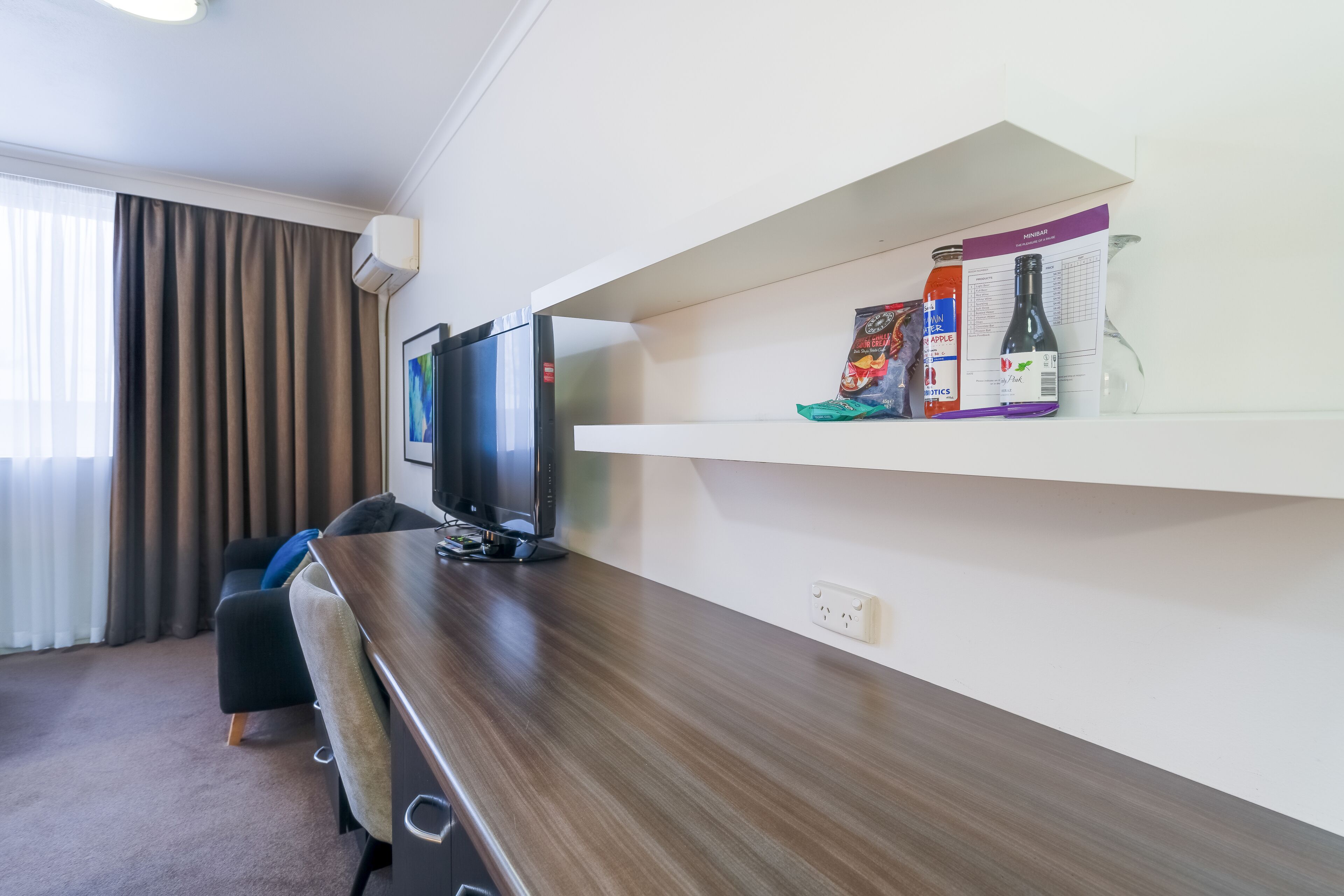 A hotel room with a wooden desk, a television, a sofa, and a shelf with items like a bottle, a box, and a glass.