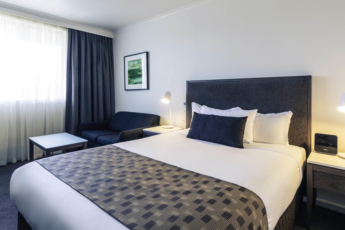 Habitación de hotel con cama, mesitas de noche, escritorio, silla, lámpara y un cuadro en la pared.