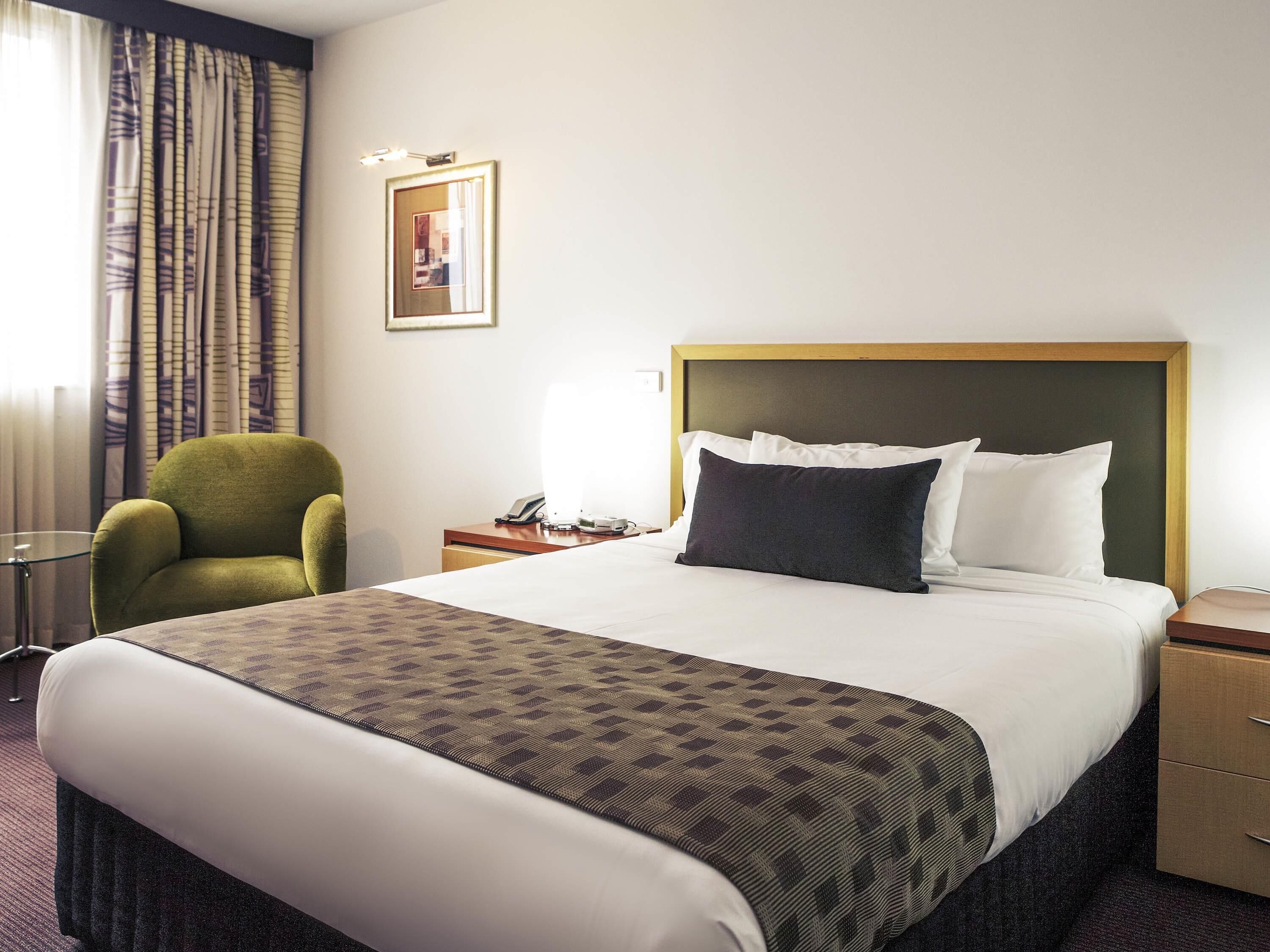 Una habitación de hotel con una cama, un sillón verde, una mesita de noche con una lámpara y un cuadro en la pared.