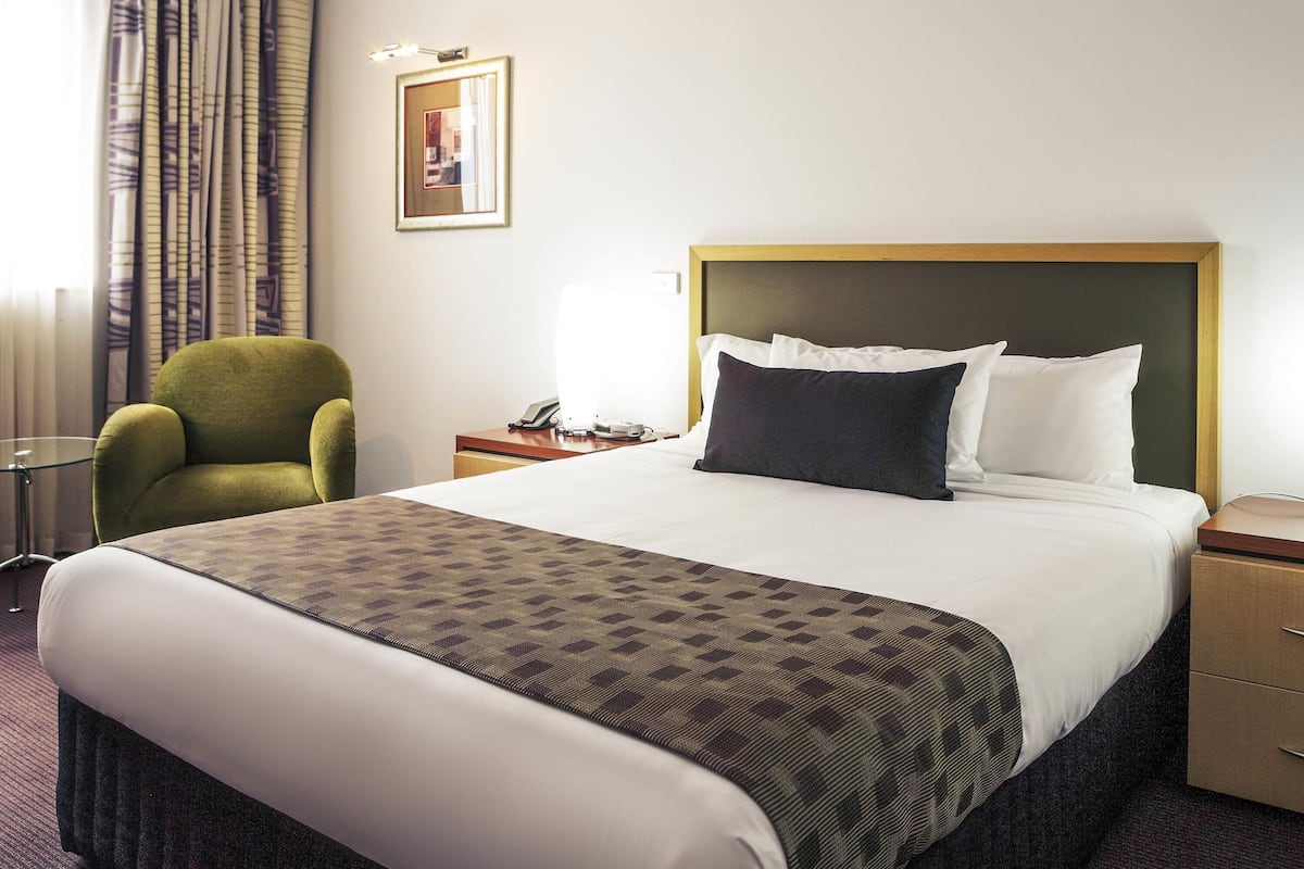 Una habitación de hotel con una cama, un sillón verde, una mesita de noche con una lámpara y un cuadro en la pared.