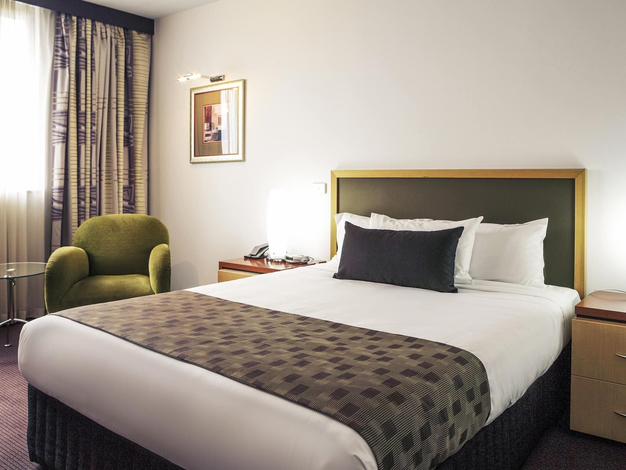 A hotel room with a bed, a green armchair, a bedside table with a lamp, and a framed picture on the wall.