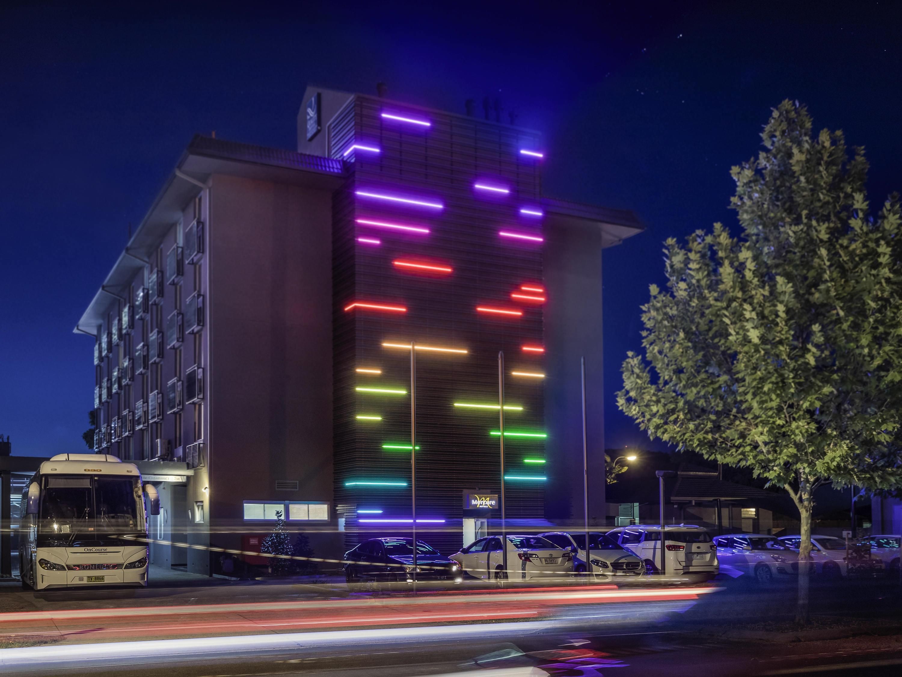 Un hotel de varios pisos con una fachada moderna, un estacionamiento con varios automóviles y un autobús detenido afuera.