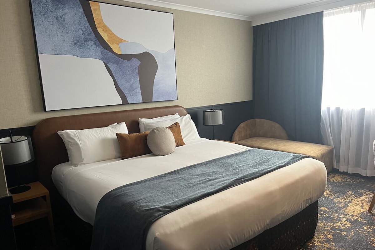 A hotel room with a large bed, a bedside table with a lamp, a chair, a large abstract painting on the wall, and a view of the outdoors through a window with curtains.