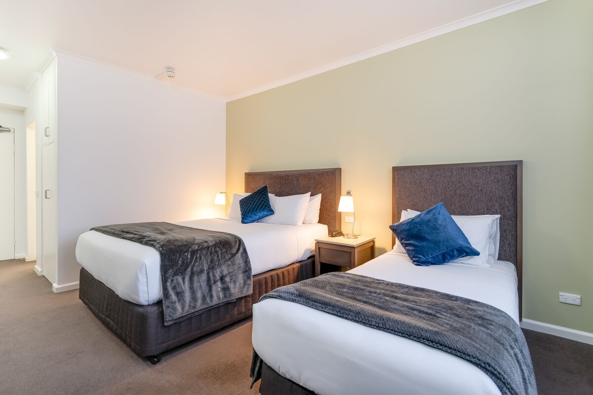 A hotel room with two beds, each with a nightstand and lamp, and a door to the left.
