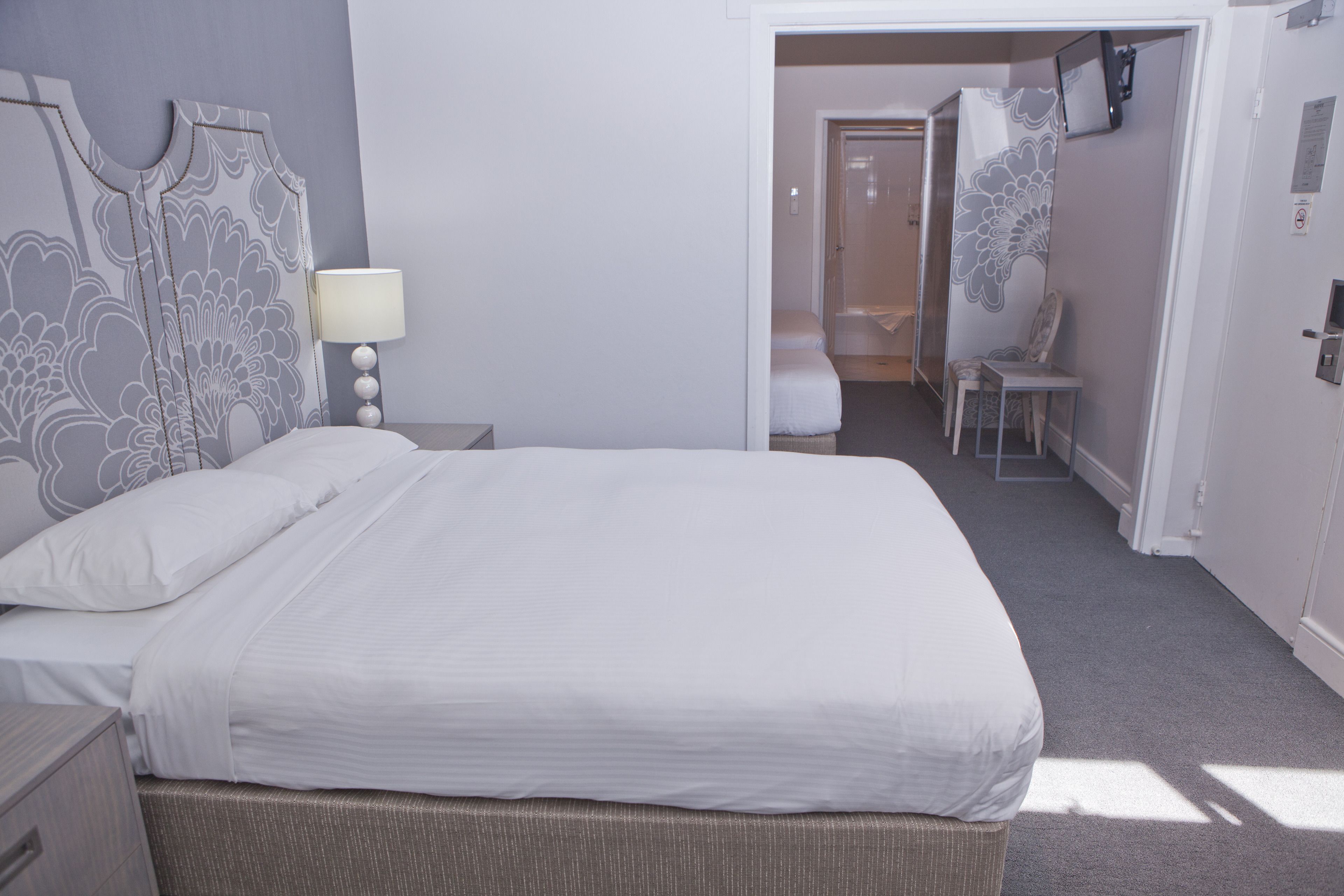 A neatly made bed with a headboard, a bedside table with a lamp, and a chair in a room with a door leading to another area.