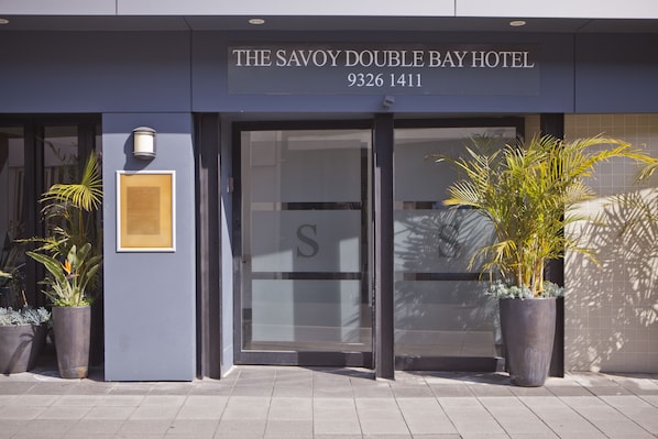 The exterior of The Savoy Double Bay Hotel with a large 'S' logo on the glass doors, a framed sign on the wall, and potted plants in front.
